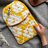 Person holiding a quilted hot water bottle cover in the vibrant Yellow Weaver print, featuring colourful birds and floral motifs on a bright yellow background, against a grey blanket.