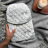 Person holding a quilted cotton hot water bottle cover in the white and grey Valley Views print, against a grey blanket with a lit candle in the background.