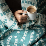Person holding a cup of tea, snuggled up under a blue-green woven cotton throw with a colourful prayer flag design.